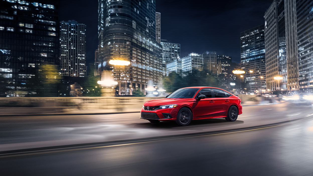 3/4 front view of greyish blue Civic Si taking a turn on a city street with lots of greenery on a sunny day.   3/4 front sideview of a red Civic Si driving on city road at night.