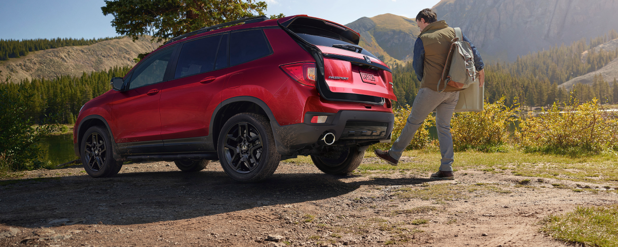 3/4 rear view of red Passport, outdoors. Man waving foot under bumper, opening trunk.