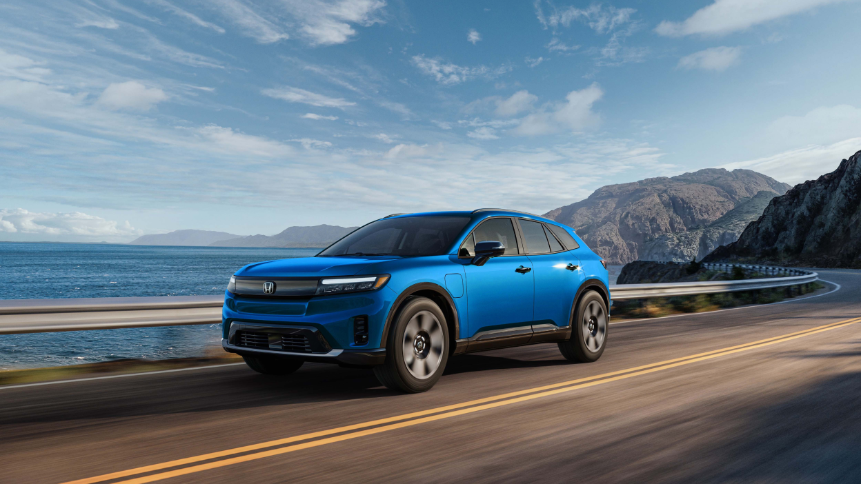 Wide 3/4 front view of blue Prologue driving on coastal highway ith mountains in the background.